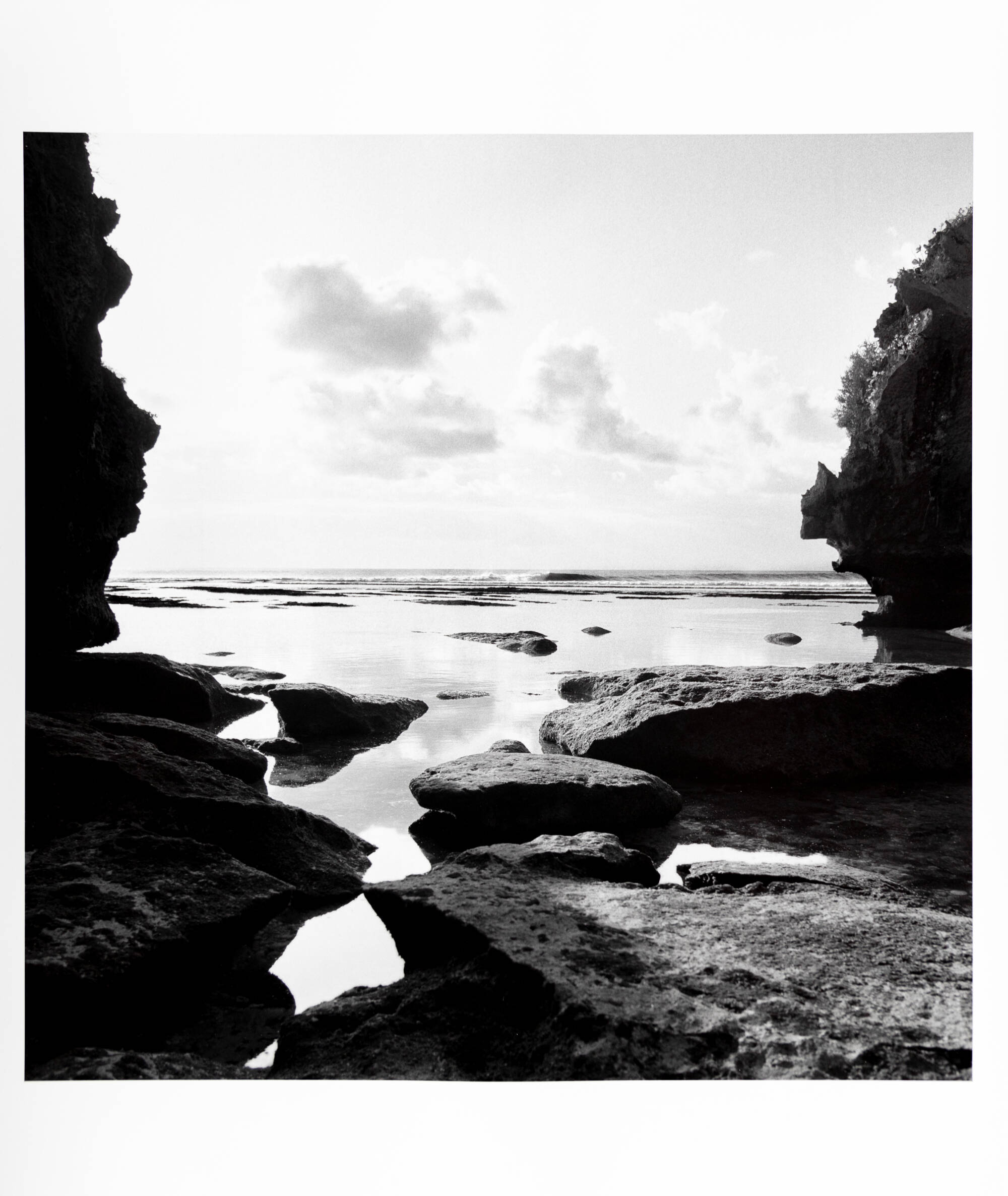 A black and white photograph of a river flowing through a group of rocks. The water is very calm.